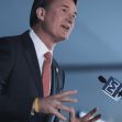 A person speaking at an event, gesturing with hands and wearing a suit and tie, with a microphone in front. A person speaking at an event, gesturing with hands and wearing a suit and tie, with a microphone in front.