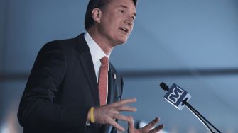 A person speaking at an event, gesturing with hands and wearing a suit and tie, with a microphone in front.