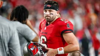 Baker Mayfield on the field, holding a football helmet while wearing a Tampa Bay Buccaneers uniform.