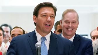 A man in a suit speaking into a microphone, surrounded by a group of people.