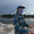 A person wearing a blue plaid shirt and a face covering operates a boat on calm waters under a cloudy sky. A person wearing a blue plaid shirt and a face covering operates a boat on calm waters under a cloudy sky.