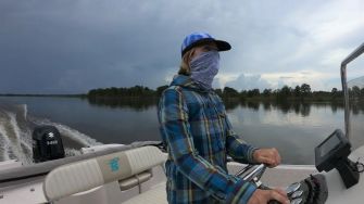 A person wearing a blue plaid shirt and a face covering operates a boat on calm waters under a cloudy sky.