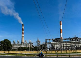 Industrial smokestacks emitting smoke under a clear blue sky, with power lines in the foreground.