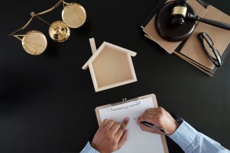 A person signing a document with a wooden house model and legal scales in the background.