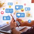 A person using a smartphone while sitting at a desk with a laptop, surrounded by various social media interaction icons and emojis. A person using a smartphone while sitting at a desk with a laptop, surrounded by various social media interaction icons and emojis.