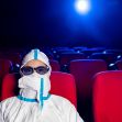 A person in a protective suit and glasses sitting alone in a movie theater with red seats and a spotlight in the background. A person in a protective suit and glasses sitting alone in a movie theater with red seats and a spotlight in the background.