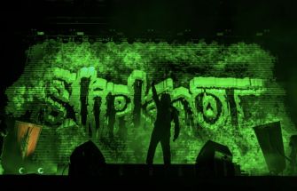 Silhouette of a musician performing on stage with a backdrop displaying the "Slipknot" logo in green lights.