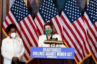 A press conference featuring lawmakers advocating for the reauthorization of the Violence Against Women Act, with flags in the background.