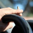 A hand holding a marijuana cigarette while gripping a car steering wheel. A hand holding a marijuana cigarette while gripping a car steering wheel.