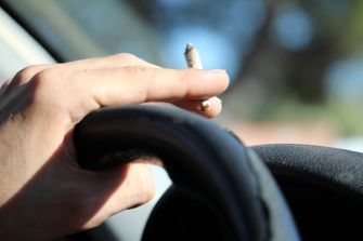 A hand holding a marijuana cigarette while gripping a car steering wheel.