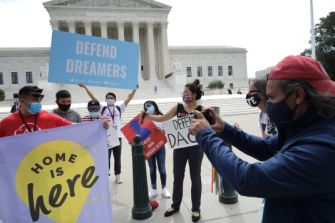A group of individuals protesting outside a government building, holding signs that read "DEFEND DREAMERS" and "DEFEND DACA," advocating for the protection of DACA recipients.