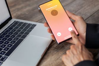 A person using a mobile phone receiving a call labeled "Marketing Corp" while sitting at a desk with a laptop.