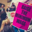 A protester holding a sign that reads "RAISE THE MINIMUM WAGE" during a demonstration. A protester holding a sign that reads "RAISE THE MINIMUM WAGE" during a demonstration.