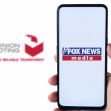A person holding a smartphone displaying the Fox News logo alongside Dominion Voting Systems branding. A person holding a smartphone displaying the Fox News logo alongside Dominion Voting Systems branding.