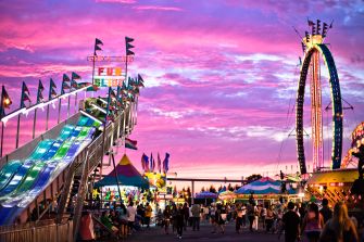 Carnival scene at sunset featuring a slide, Ferris wheel, and festival-goers.