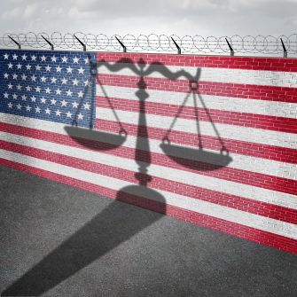 A shadow of scales of justice cast on a wall painted with the American flag, with barbed wire along the top.