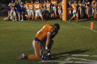 A football player kneeling in prayer on the field after a game, with teammates in the background.
