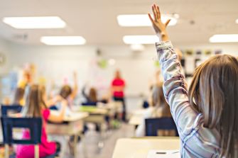A classroom setting with students raising their hands, focusing on a teacher in the background.