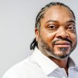 A man with braided hair and a beard, wearing a white shirt, looks into the camera with a subtle smile against a neutral background. A man with braided hair and a beard, wearing a white shirt, looks into the camera with a subtle smile against a neutral background.