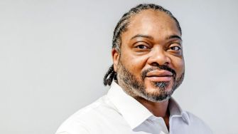 A man with braided hair and a beard, wearing a white shirt, looks into the camera with a subtle smile against a neutral background.
