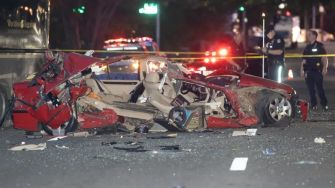 A severely damaged red car on a street after a deadly crash, with police officers and emergency vehicles in the background.