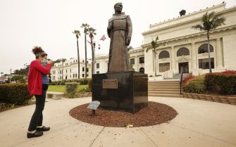 Statue of Father Junipero Serra outside a historic building, with a woman taking a photograph.