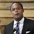 Man speaking at a podium with a microphone, wearing a suit and tie, with an American flag in the background. Man speaking at a podium with a microphone, wearing a suit and tie, with an American flag in the background.