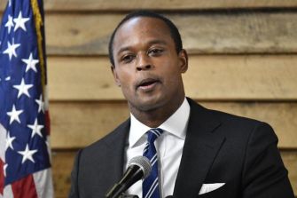 Man speaking at a podium with a microphone, wearing a suit and tie, with an American flag in the background.