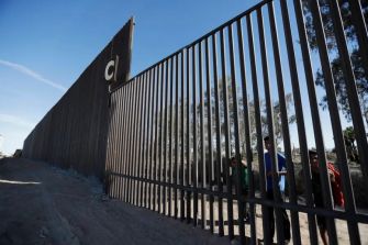 Children looking through gaps in a section of the border wall.