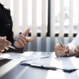 Two individuals discussing and signing legal documents at a table, with sunlight casting shadows through the blinds in the background. Two individuals discussing and signing legal documents at a table, with sunlight casting shadows through the blinds in the background.