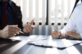 Two individuals discussing and signing legal documents at a table, with sunlight casting shadows through the blinds in the background.