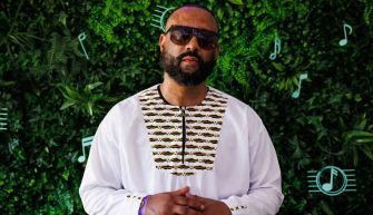 A man in sunglasses and a white traditional outfit standing in front of a green foliage backdrop.