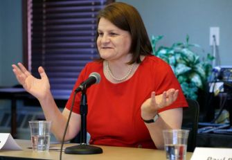 A woman speaking at a podium with a microphone, gesturing with her hands during a discussion, surrounded by glasses of water and plants.
