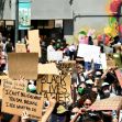 A large crowd of protesters holding signs advocating for racial justice, with a mural of George Floyd in the background. A large crowd of protesters holding signs advocating for racial justice, with a mural of George Floyd in the background.