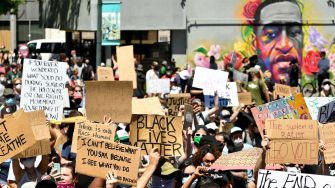 A large crowd of protesters holding signs advocating for racial justice, with a mural of George Floyd in the background.