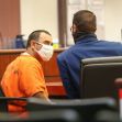 Two individuals in a courtroom; one is wearing an orange prison jumpsuit and a mask, while the other is in formal attire, also wearing a mask. Two individuals in a courtroom; one is wearing an orange prison jumpsuit and a mask, while the other is in formal attire, also wearing a mask.