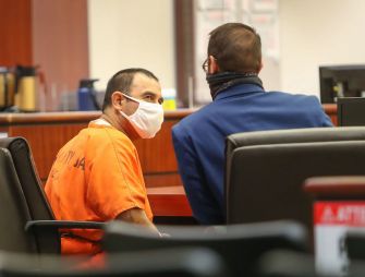 Two individuals in a courtroom; one is wearing an orange prison jumpsuit and a mask, while the other is in formal attire, also wearing a mask.