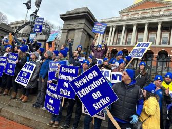 A group of educators holding signs in support of the Newton Teachers Association strike outside a government building.