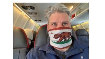 A man wearing a California flag face mask while seated on an airplane.
