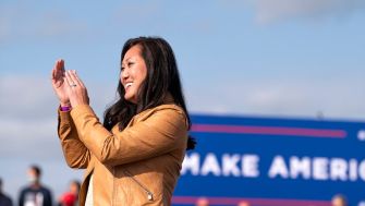 A woman claps while standing outdoors, with a "Make America Great Again" banner visible in the background.