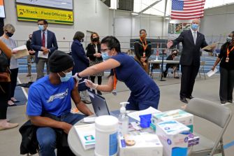 A healthcare worker administering a COVID-19 vaccine to an individual in a vaccination center, with officials observing in the background.