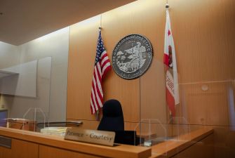 Image of a courtroom with flags and a seal of California, featuring a judge's bench and signage that reads "Patience - Courtesy."