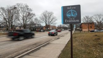Welcome sign for the city of Evanston, Illinois, alongside a street with passing cars and bare trees.