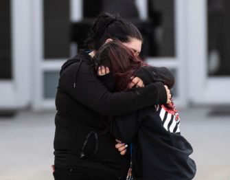 Two individuals embrace in a moment of grief outside a school, reflecting the emotional aftermath of a tragic incident.