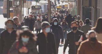 A busy street scene showing a diverse crowd of people wearing masks, reflecting ongoing public health measures during the COVID-19 pandemic.