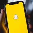 A person holding a smartphone displaying the Snapchat logo on a yellow background. A person holding a smartphone displaying the Snapchat logo on a yellow background.