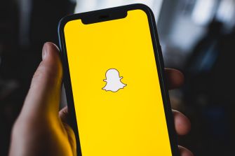 A person holding a smartphone displaying the Snapchat logo on a yellow background.