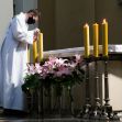 A masked individual in church attire adjusts candles next to a floral arrangement on an altar. A masked individual in church attire adjusts candles next to a floral arrangement on an altar.