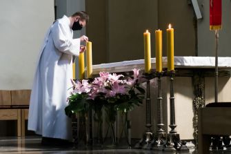 A masked individual in church attire adjusts candles next to a floral arrangement on an altar.