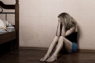 A woman sitting on the floor, holding her head in distress, with a bed in the background.